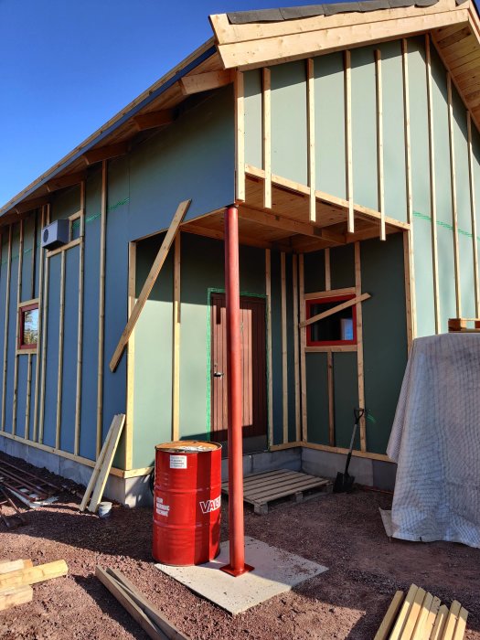 Hus under renovering visar en provmonterad stolpe, träpanel, och byggmaterial runt omkring.