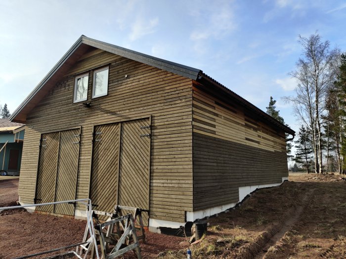 Nybyggt hus med delvis järnvitriolerad träpanel, stora stängda garageportar och stegar framför fasaden.