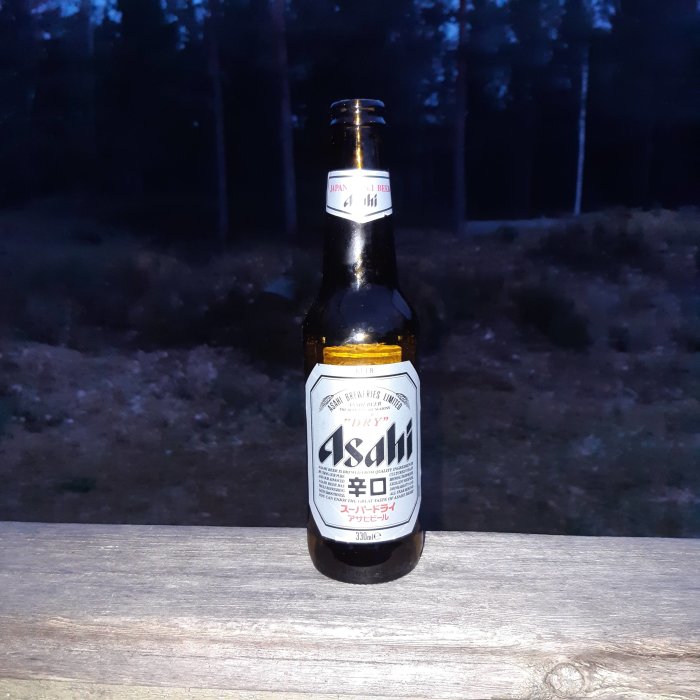 En flaska Asahi dry beer står på ett träbord med en skymningsskog i bakgrunden.