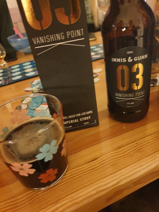 En ölflaska och ett glas fyllt med mörk öl bredvid en förpackning för "Vanishing Point" på ett träbord.