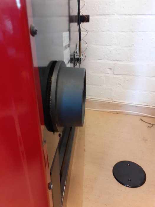 Sidovy av en röd spis med svart stos ansluten till väggen och ett vitt locket på golvet.