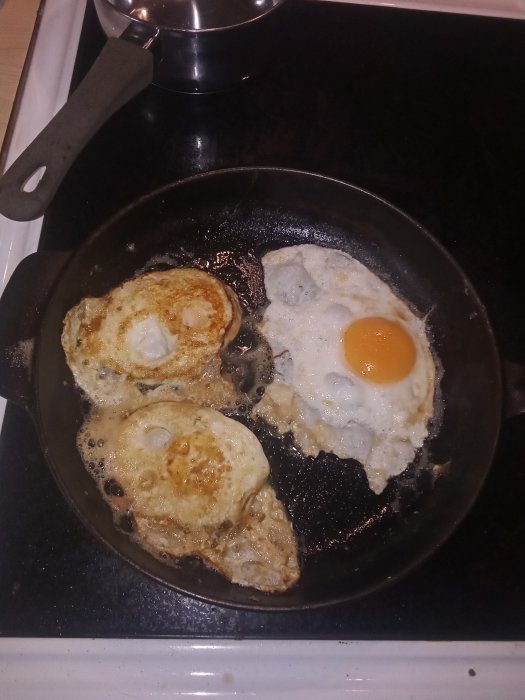 Stekta ägg i olika stadier av tillagning på en svart stekpanna ovanpå en spis.