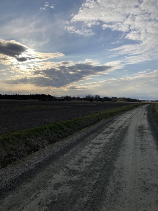 Grusväg genom åkerlandskap med molnig himmel och solstrålar som lyser igenom molnen.