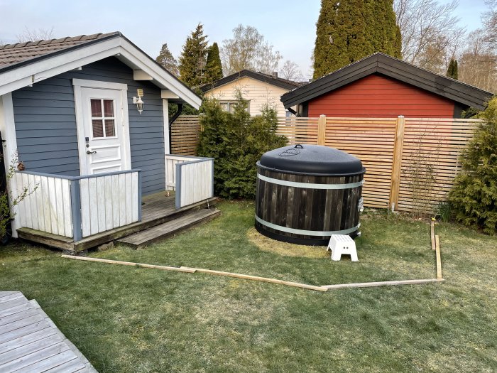 Uppställd badtunna bredvid litet blått skjul, kartong på gräsmatta markerar kaminplacering, träpinnar utlagda för ny altan.