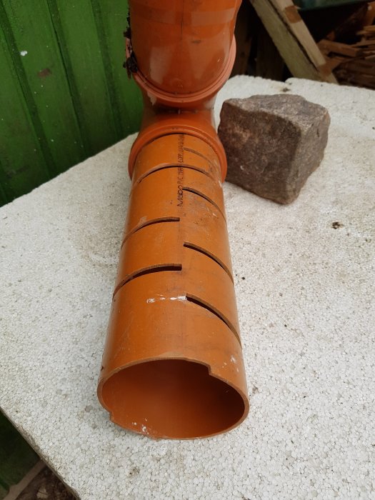 Orange avloppsrör med sågade spår och en sten som tyngd på en vit isoleringsplatta.