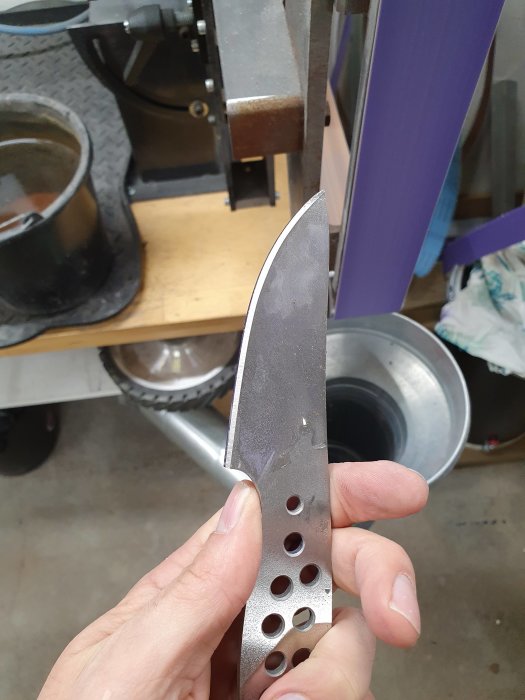 Hand håller ett knivblad med slipade kanter och hålmönster under arbete i en verkstad.