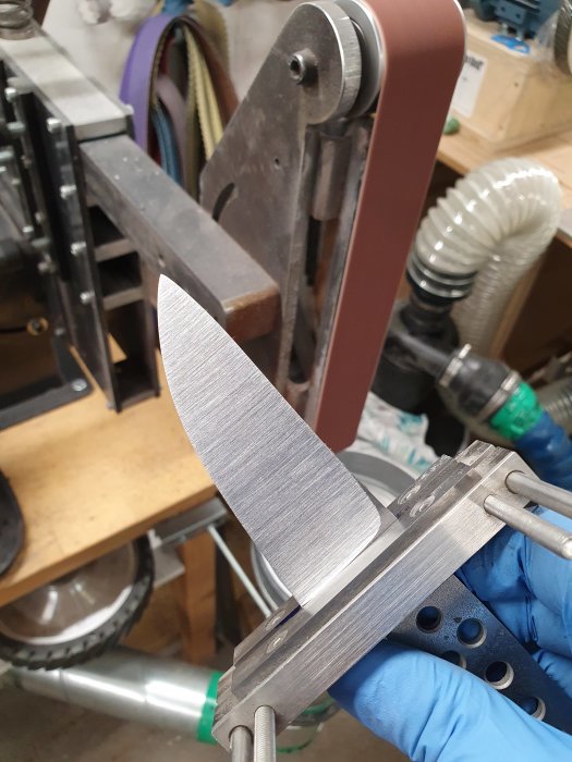 Knivblad som slipas med ett 60-korns slipband på en slipmaskin i en verkstad.