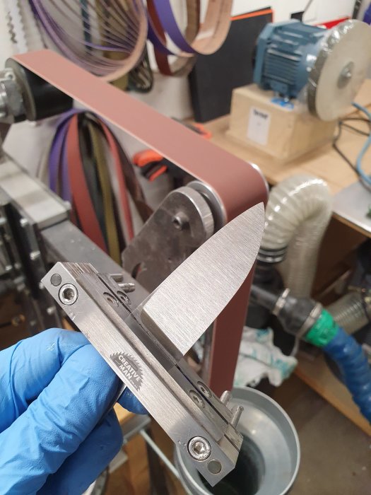 Hantverkare slipar knivblad med slipmaskin och använder olika slipband för finish.