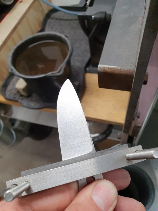Hand håller nyslipat knivblad nära bandslip, visar arbete med slipning på 120 korn.
