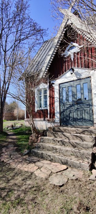 Ett rött hus med vita knutar och en stenlagd gång framför trappan, omgivet av bar trädgrenar och blå himmel.