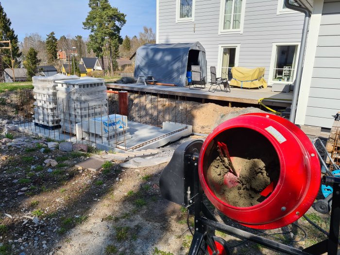 Pågående poolbygge nära grått hus med byggmaterial och verktyg, trappsteg under konstruktion, cementblandare i förgrunden.