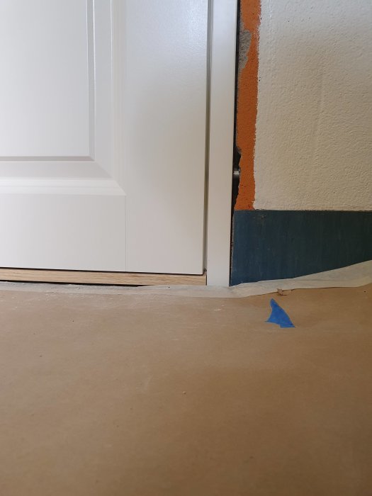 Nederdelen av en vit innerdörr som hänger snett i karmen intill en ojämnt målad beige och blå vägg.