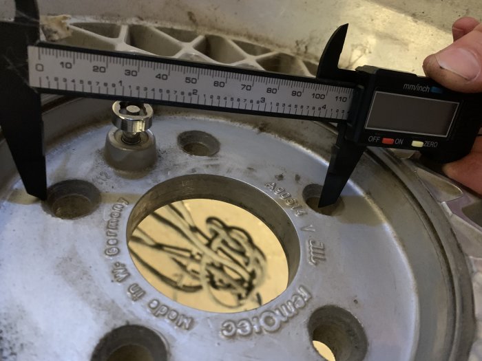 Hand använder en digital skjutmått för att mäta avståndet mellan hjulbultar på en bilfälg med text och logo.