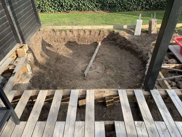 Uppgrävt hål för en framtida pool sedd från en trätrall, omgivet av gräsmatta och några betongplintar.
