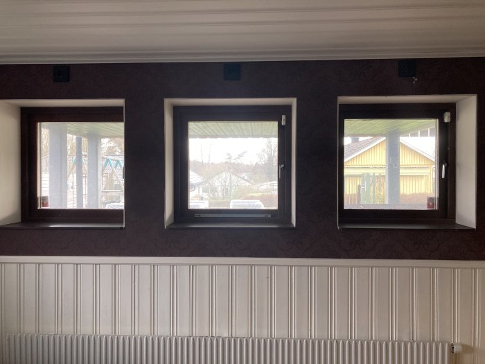Vägg med tre vita fönster, brun tapet ovanför och vit bröstpanel samt en radiator under fönstren.