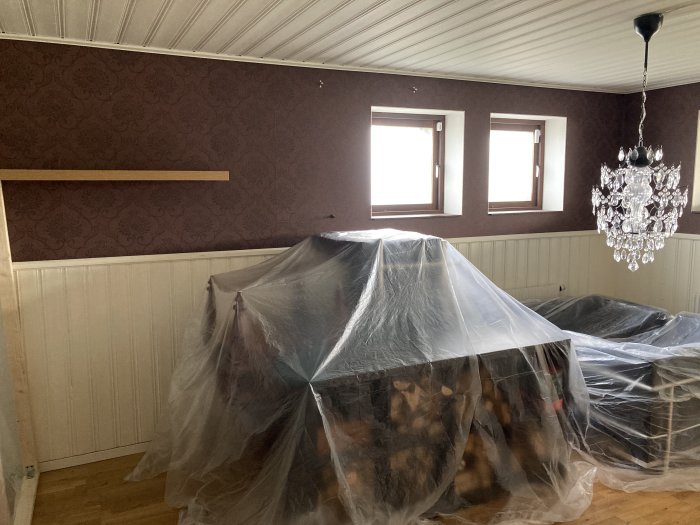 Renoveringsrum med bruna väggar, vit bröstpanel, fönster och möbler täckta med plast samt en kristallkrona.