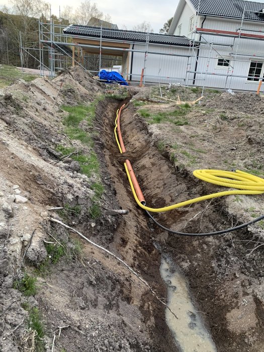 Grävt dike mellan hus och garage med rör och slangar lagda i förberedelse för att fylla igen och skapa gräsmatta.