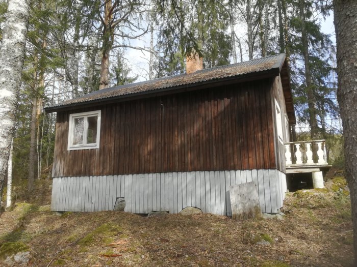 En träfasad på en sommarstuga med ett litet fönster, omgiven av träd och med en veranda på sidan.