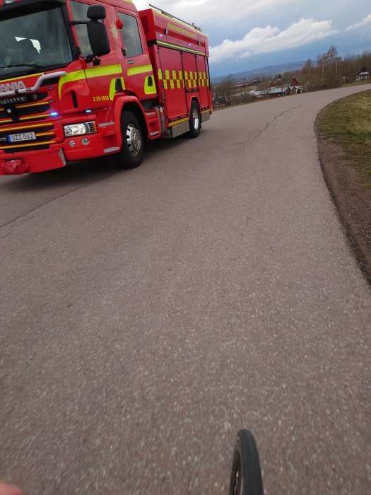 Röd brandbil i rörelse på väg, del av cykel eller scooter synlig i förgrunden.