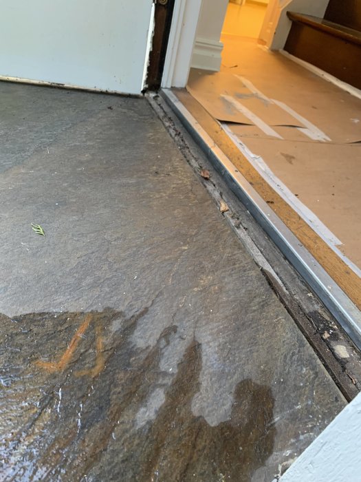 Ytterdörrs tröskel med skadat trä och saknat bleck, samt en våt trappa som lutar mot dörren.