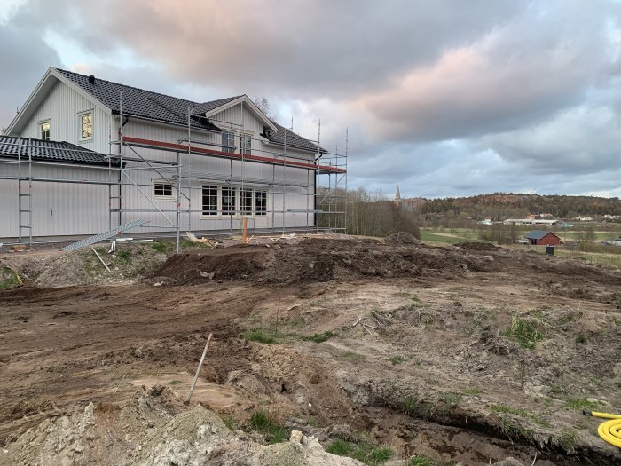 Hus under renovering med byggställningar och ojämna jordhögar i förgrunden från grovplanering.