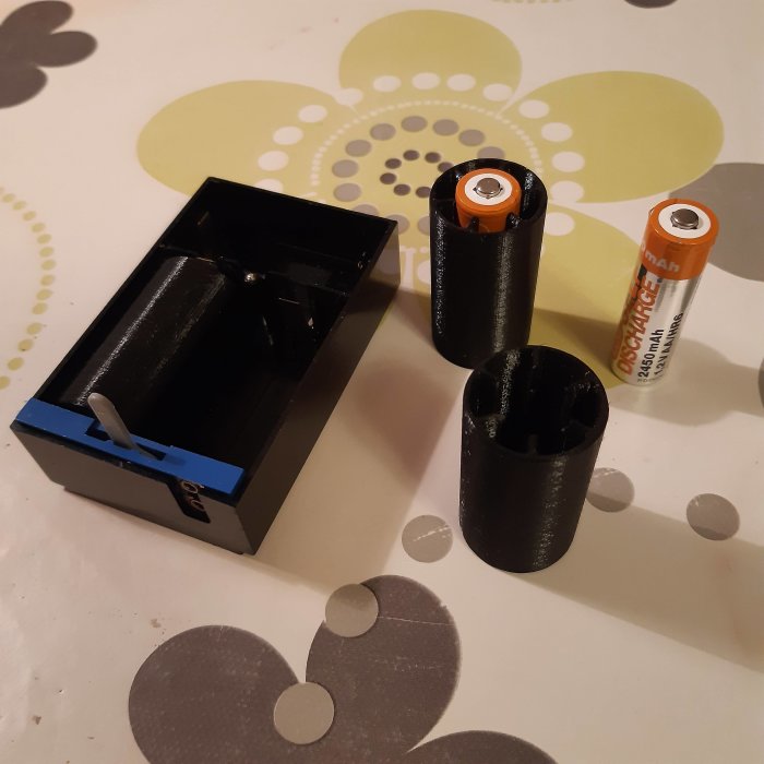 3D-utskrivna batterihållare bredvid AA-batterier på ett prickigt bord.