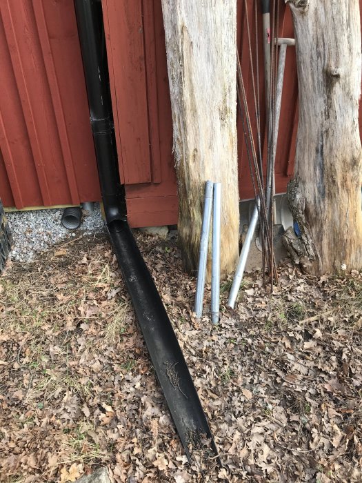 Svart stuprör lutat mot röd stuga och vedträn bredvid metallstänger på mark täckt med löv.