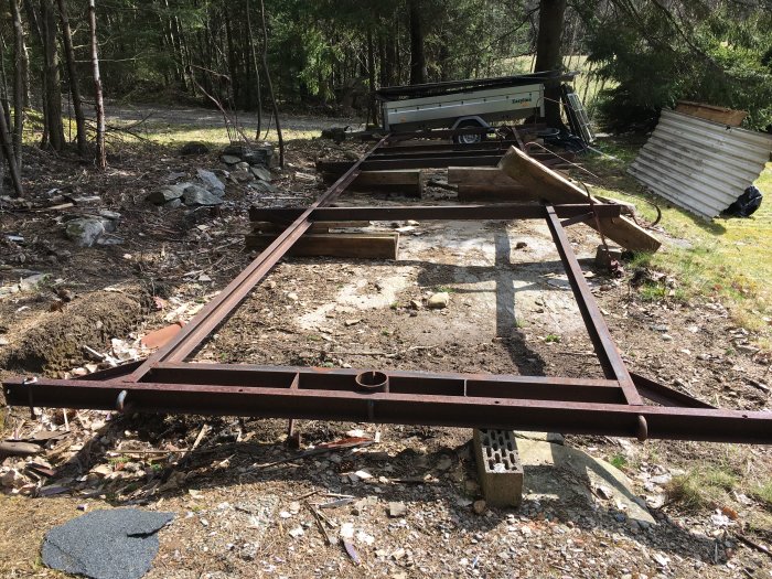 Rostigt metallunderrede i skogsmiljö, med böj på ena kortsidan och byggmaterial i bakgrunden.