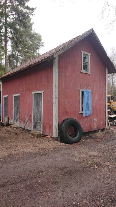 Gamla röda uthusbyggnaden som ska rivas med slitna dörrar och fönster, mot grusväg med träd och grävmaskin i bakgrunden.