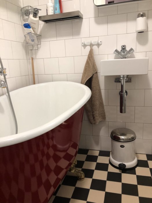 Litet badrum med röd fotad gjutjärnsbadkar, vit vägghängd handfat, och vit-svart rutigt golv.
