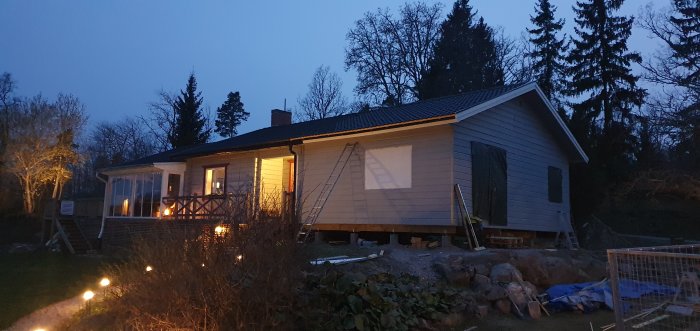 Kvällsbild av hus under renovering med tända lampor och stege vid fasaden.