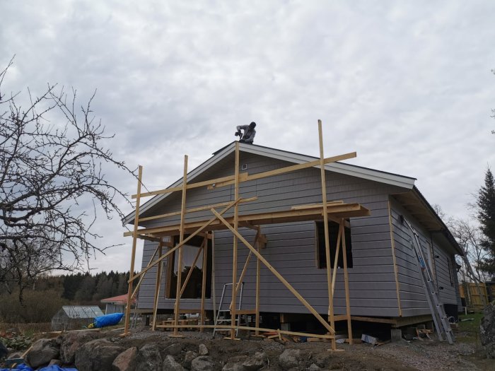 Person arbetar på taket av ett nästan färdigt trähus, med byggmaterial och ställningar synliga.