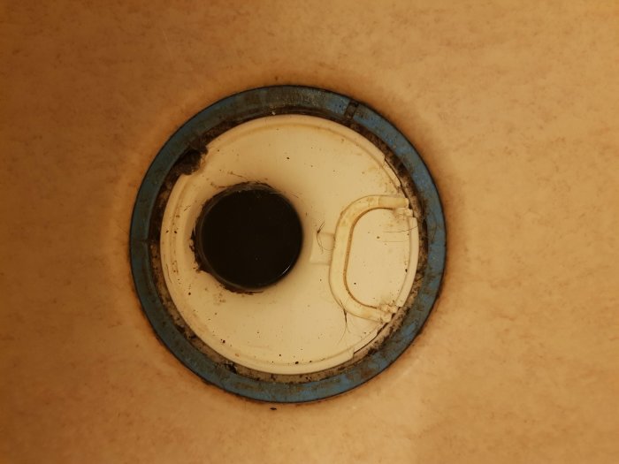 Närbild av en äldre golvbrunn i ett badrum, med synliga tecken på slitage och smutsansamlingar.