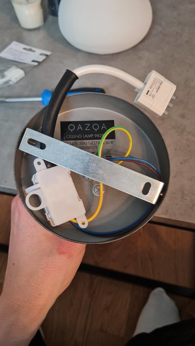 Hand håller en taklampa med installationsplatta och elkabel framför en takkrok.