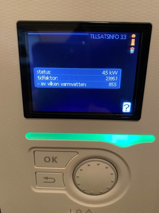 Display på värmepump med värden för status, tidfaktor och varmvatten samt kontrollpanel med grönt ljus.