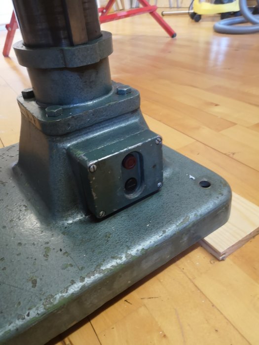 Närbild av en grå maskinpelare i metall med en brytare monterad nere vid foten, mot en trägolvsbakgrund.