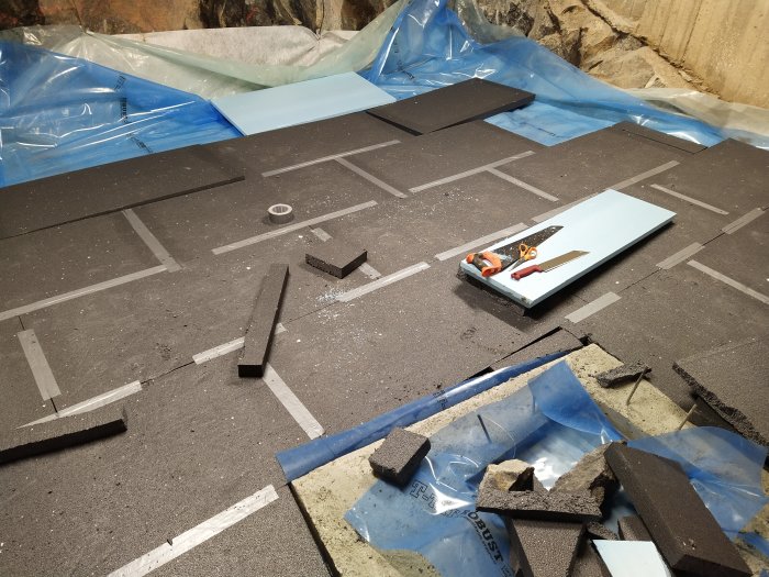 Ojämnt lagda cellplastskivor på betonggolv med tejpmarkeringar och byggmaterial spridda omkring.