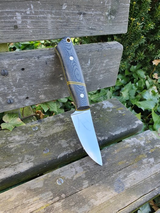 Kniv med format och slipat blått skaft vilar på en grå träbänk, polerat blad reflekterar ljus.