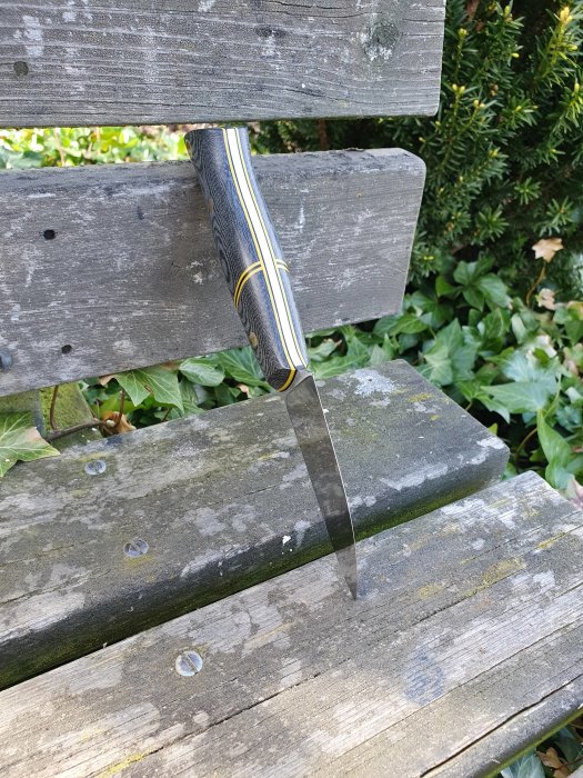 Välslipad kniv med mönstrat skaft som sticker igenom två träplankor.