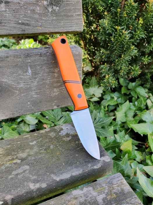Handgjord kniv med orange skaft, finslipat och polerat blad, mot en grå träbänk och grönska.
