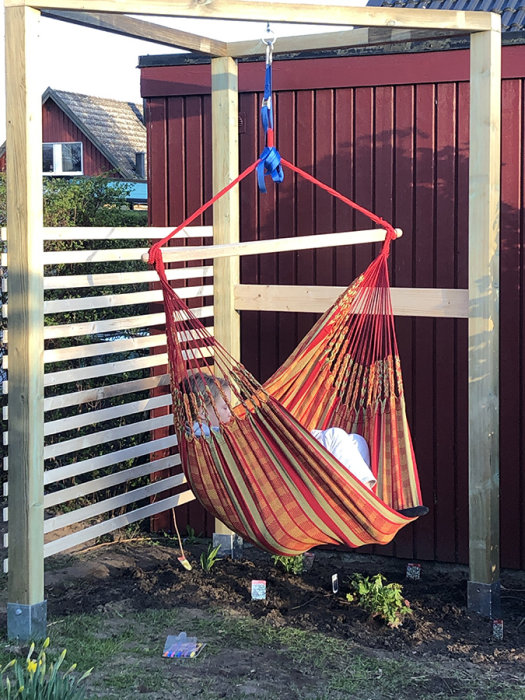 En röd och orange randig hängstol hängande i en enkel träställning vid ett rött staket i en trädgård.