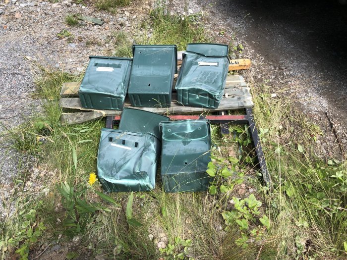 Deformerade gröna brevlådor hopslängda på marken bredvid en grusväg, tecken på skada efter olycka.