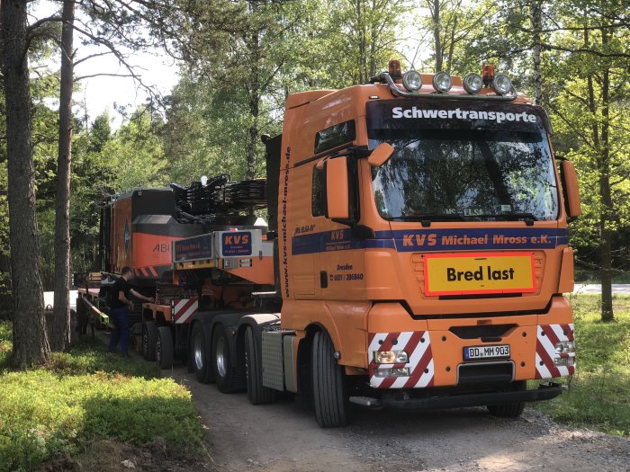 Orange lastbil märkt "Schwertransporte" transporterar en stor pålkran, skyltad "Bred last".