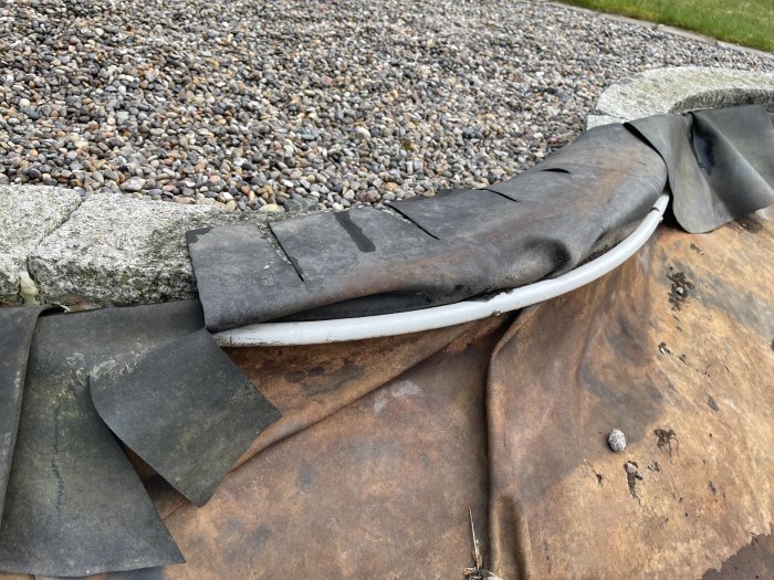 Veckig gummiduk fastsatt vid granitkantsten med rör i en trädgårdsdamm, omgiven av grus.