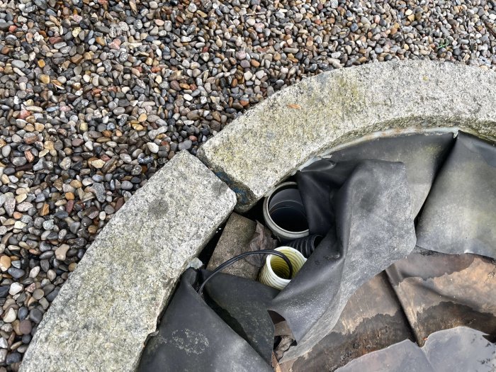 Granitkant med en veckig gummiduk och rör synliga vid en damms kant, omgivna av grus.
