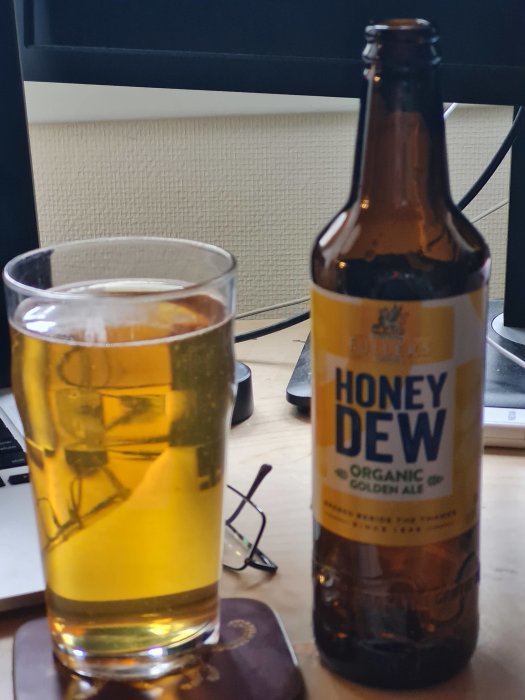 Glas med blekgul öl bredvid en flaska Honey Dew Organic Golden Ale på ett skrivbord.