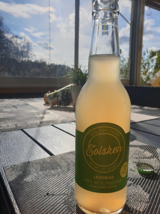 Flaska med citronlemonad på ett bord i soligt uterum, med suddig bakgrund av träd och himmel.