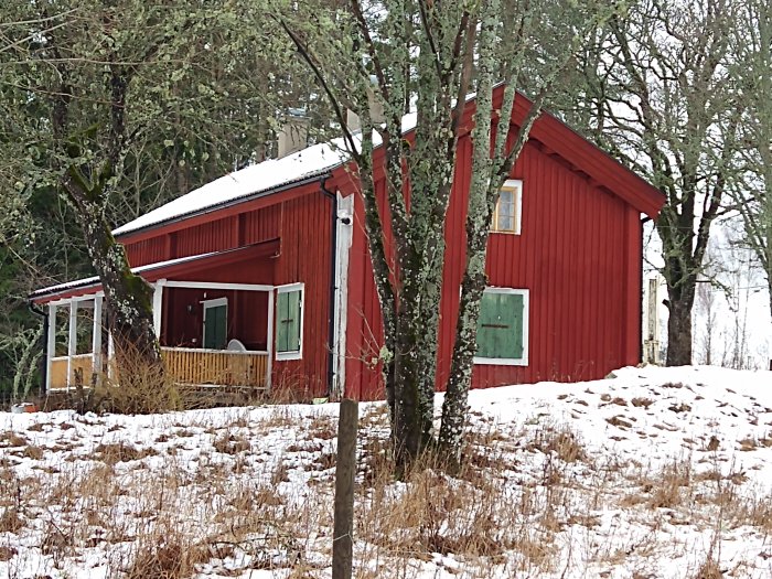 Rött litet hus med snö på taket omgivet av snötäckt mark och lövträd, i Örebro län.