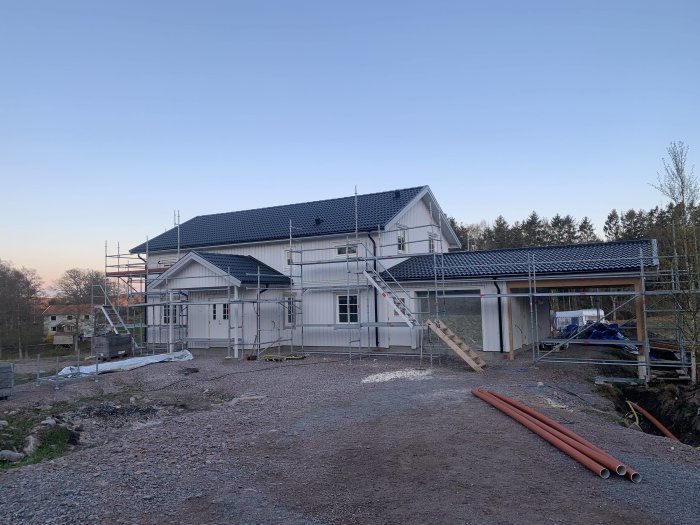 Ett hus under renovering med byggställningar och byggmaterial synliga på gården i skymningen.