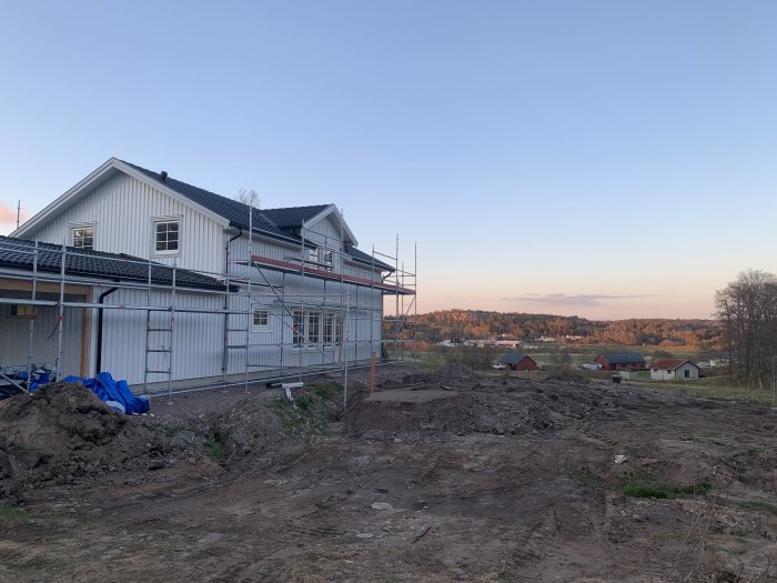 Hus under renovering med byggnadsställningar och en röjd mark framför vid skymning.